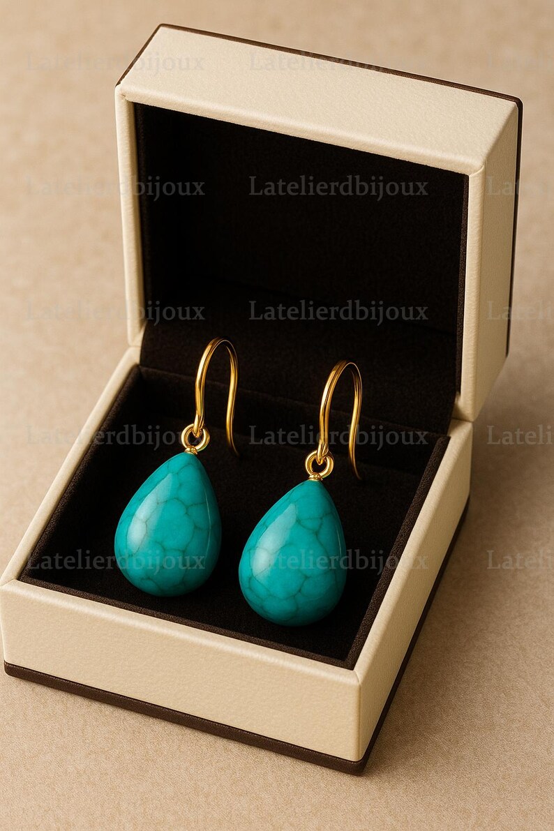 Puede incluir: Un par de pendientes turquesa en forma de l&aacute;grima con ganchos dorados, presentados en una caja de joyer&iacute;a beige con interior negro. Los pendientes tienen una superficie texturizada. El texto "Latelierdbijoux" es visible.