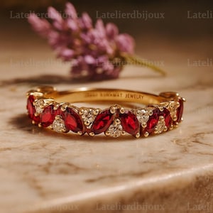 Puede incluir: Un anillo de banda dorado con piedras rojas en forma de marquesa y pequeñas piedras blancas alternadas. El anillo está sobre una superficie de mármol con una flor morada borrosa en el fondo. El texto "NOWAWAT JEWELRY" está grabado en el anillo.