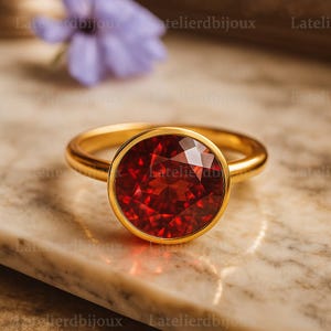 以下が含まれることがあります： 大きな丸い赤い宝石が付いた金の指輪。指輪はシンプルなベゼルセッティングで、バンドは滑らかで磨かれています。背景はぼやけた大理石の表面です。