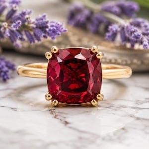 Può includere: Un anello in oro con una grande pietra preziosa rosso intenso, taglio a cuscino, incastonata in una montatura a quattro griffe. La fascia è in oro liscio e lucido, offrendo un netto contrasto con la pietra rossa sfaccettata e vivace al centro.