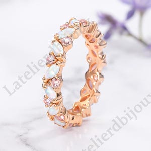 May include: Rose gold ring featuring alternating opal and pink gemstone settings. The ring's design includes oval-shaped opals and round pink gemstones, creating a continuous band of color and sparkle. The ring is set on a white marble surface.