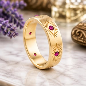 May include: A gold ring featuring an engraved eye motif and small red gemstones. The ring has a smooth, polished finish. The background is blurred, with purple flowers visible.