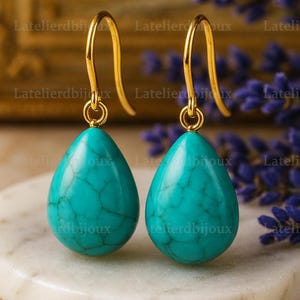 Puede incluir: Un par de pendientes colgantes dorados con piedras turquesas en forma de l&aacute;grima. Los pendientes se exhiben sobre una superficie de m&aacute;rmol blanco, con un fondo borroso de flores moradas y detalles dorados.