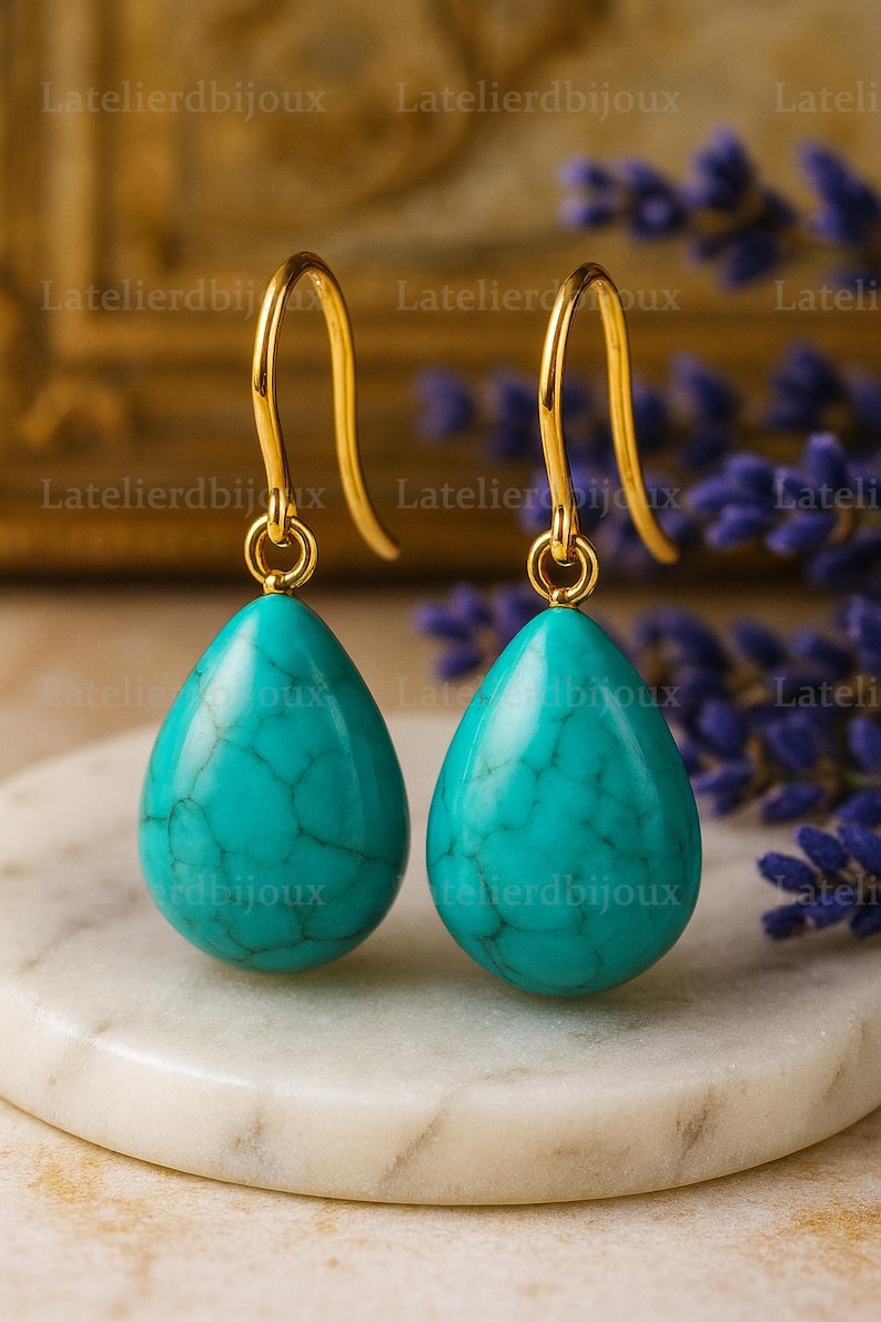 Puede incluir: Un par de pendientes colgantes dorados con piedras turquesas en forma de l&aacute;grima. Los pendientes se exhiben sobre una superficie de m&aacute;rmol blanco, con un fondo borroso de flores moradas y detalles dorados.