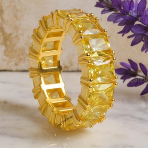 Könnte beinhalten: Ein goldfarbener Ring mit einem Band aus gelben, rechteckigen Edelsteinen. Der Ring ist vor einem Marmorhintergrund mit lila Blumen platziert. Der Ring ist ein Voll-Eternity-Band.