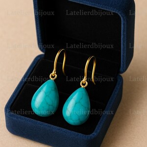 Puede incluir: Un par de pendientes turquesa en forma de l&aacute;grima con ganchos dorados, presentados en una caja de joyer&iacute;a de terciopelo azul oscuro. Los pendientes tienen un aspecto veteado. El fondo es neutro.