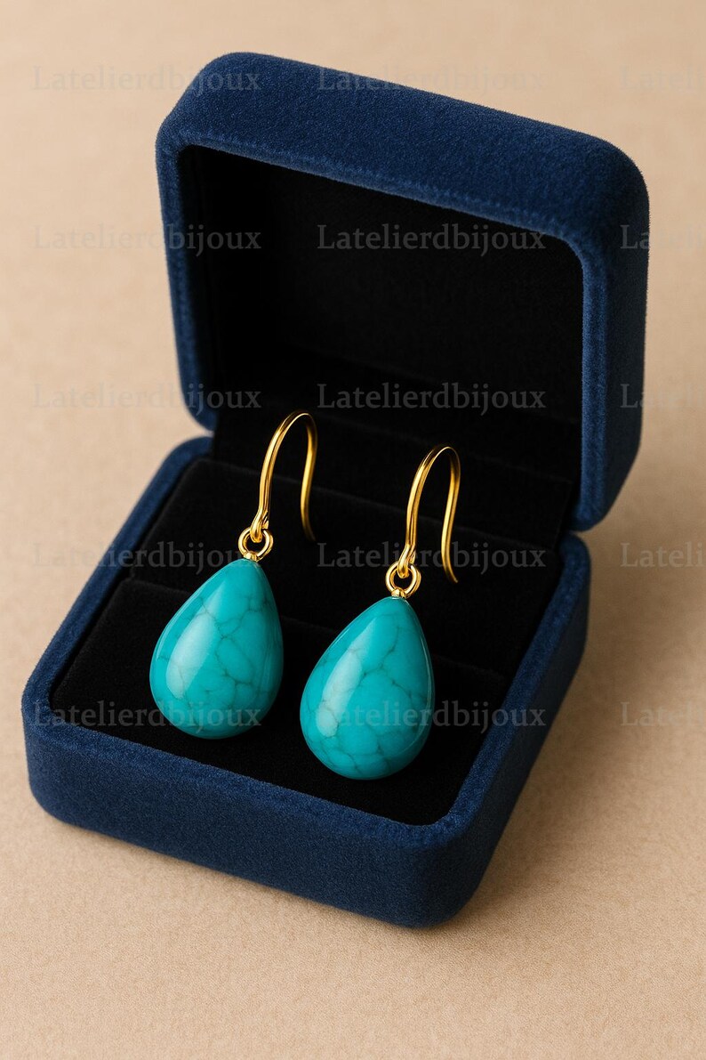 Puede incluir: Un par de pendientes turquesa en forma de l&aacute;grima con ganchos dorados, presentados en una caja de joyer&iacute;a de terciopelo azul oscuro. Los pendientes tienen un aspecto veteado. El fondo es neutro.