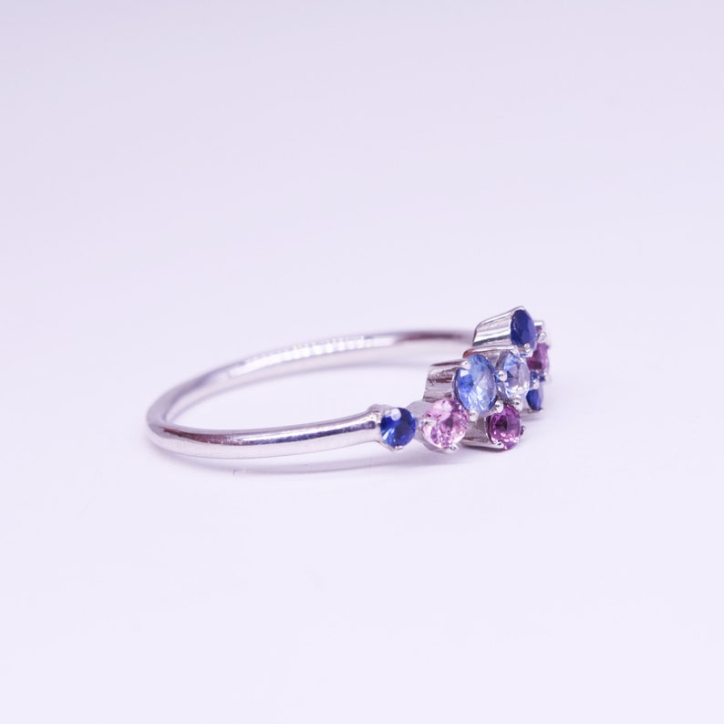 Puede incluir: Un anillo de plata con un grupo de peque&ntilde;as piedras preciosas de colores. El anillo presenta una banda delicada y est&aacute; adornado con piedras azules, moradas y rosas, creando un dise&ntilde;o brillante y elegante. Las piedras est&aacute;n dispuestas en un patr&oacute;n asim&eacute;trico &uacute;nico.