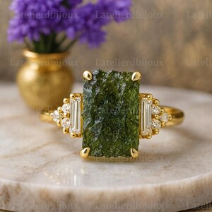 Puede incluir: Un anillo de oro con una gran piedra preciosa verde rectangular y texturizada en el centro. El anillo está adornado con piedras preciosas más pequeñas, claras, rectangulares y redondas. El anillo está sobre un fondo borroso. Es una joya.