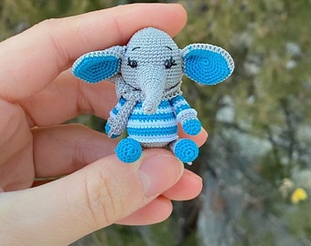 May include: A small gray crocheted elephant wearing a blue and white striped shirt. The elephant is sitting in a hand with a blurry background of green trees. The text "MINIATURE ELEPHANT" is written above the elephant.