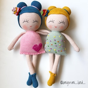 May include: Two crocheted dolls with colourful yarn hair and dresses. One doll has a pink dress with a heart on the front and the other has a green dress with colourful dots. The dolls have closed eyes and are sitting together.