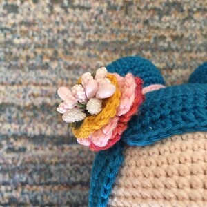 May include: A close-up of a crocheted hat with a colorful flower detail. The hat is made of blue, pink, and yellow yarn. The flower is made of pink and white beads.