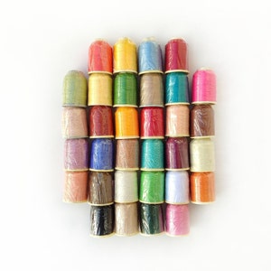 May include: A collection of 28 spools of thread in various colours, including red, orange, yellow, green, blue, purple, pink, brown, and beige. The spools are arranged in a semi-circle shape.