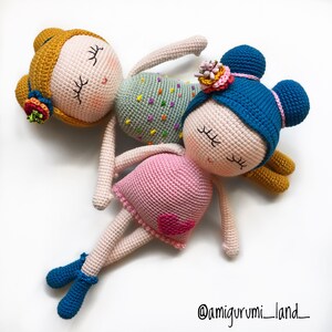 May include: Two crocheted dolls with closed eyes, one with a pink dress and blue hair, the other with a green dress and yellow hair. Both dolls have embroidered facial features and are lying on a white background. The caption '@amigurumi_land_' is visible in the bottom right corner.