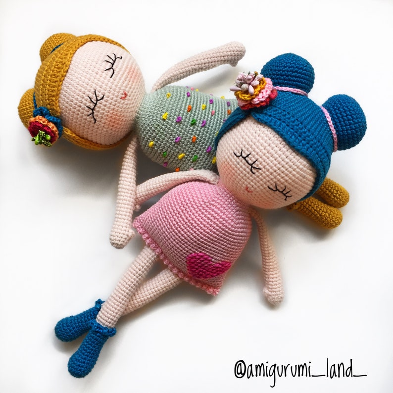 May include: Two crocheted dolls with closed eyes, one with a pink dress and blue hair, the other with a green dress and yellow hair. Both dolls have embroidered facial features and are lying on a white background. The caption '@amigurumi_land_' is visible in the bottom right corner.