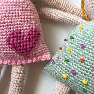 May include: Two crocheted dolls, one with a pink dress and a pink heart on the front, and the other with a light green dress with colorful sprinkles.