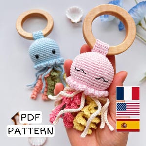 以下が含まれることがあります： 青とピンクの2つの編みぐるみのクラゲのおもちゃ。それぞれ木製のリングに付いています。クラゲはピンク、黄色、緑の色合いのカラフルな触手を持っています。画像には「PDF PATTERN」というテキストも含まれています。