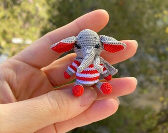 May include: A small gray crocheted elephant wearing a red and white striped shirt. The elephant has black eyes and red ears.