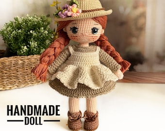 Puede incluir: Muñeca de crochet hecha a mano con cabello castaño, un vestido marrón y beige y un sombrero de paja con flores. La muñeca lleva zapatos marrones y está de pie sobre una superficie de madera. El texto "HANDMADE DOLL" está escrito debajo de la muñeca.