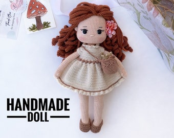 May include: Handmade crocheted doll with brown hair, a beige dress, and brown shoes. The doll is holding a small brown basket with flowers. The doll is on a white background with the text "HANDMADE DOLL" below it.