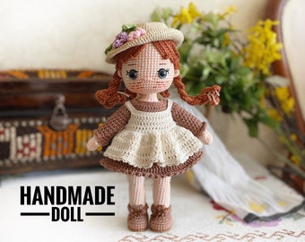 Puede incluir: Muñeca de crochet hecha a mano con cabello castaño, un vestido blanco y un sombrero marrón con flores rosas. La muñeca está de pie sobre una superficie blanca con un patrón floral.