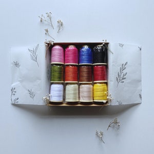 May include: A box of 12 spools of embroidery floss in various colors, including white, yellow, orange, red, pink, blue, green, and brown. The spools are arranged in a grid pattern in a cardboard box.