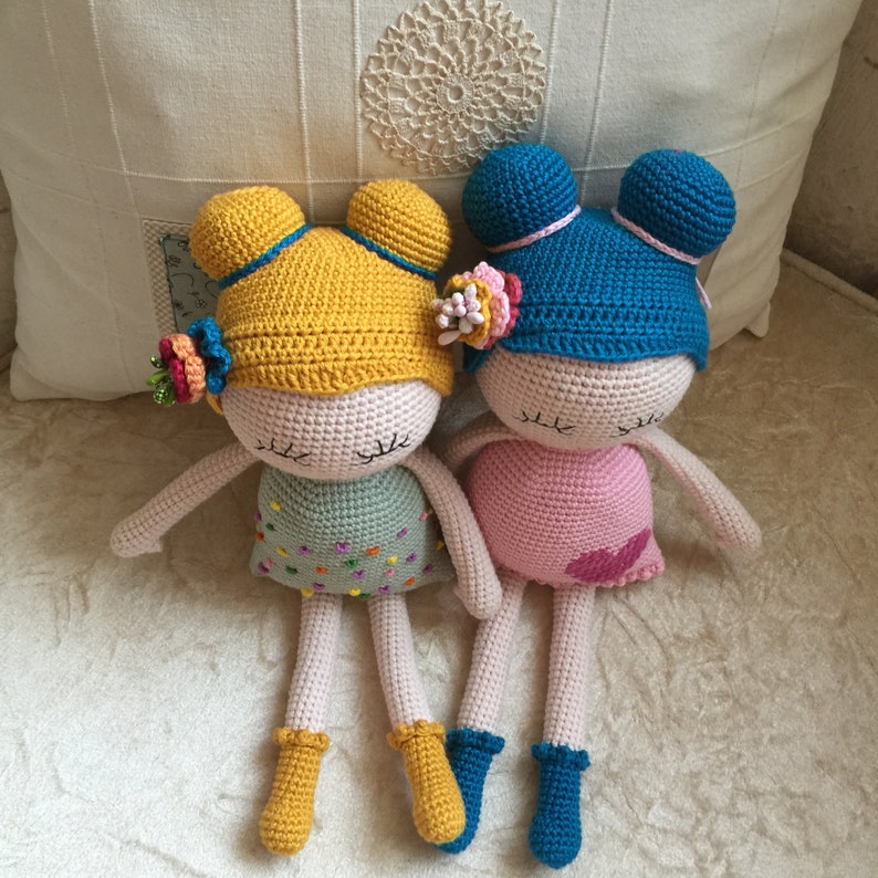 May include: Two crocheted dolls with closed eyes. One doll has a yellow hat and a green dress with colorful dots. The other doll has a blue hat and a pink dress with a pink heart.