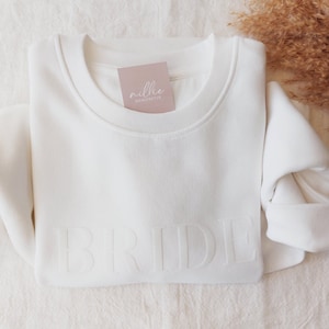 Könnte beinhalten: Weißer Pullover mit dem Wort "BRIDE" auf der Vorderseite geprägt. Der Pullover hat ein kleines Etikett auf der Vorderseite, auf dem "millie manufaktur" steht.