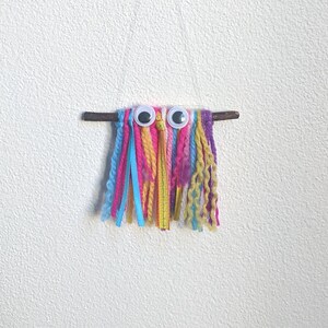 May include: A colourful yarn and ribbon craft with googly eyes attached to a small branch. The yarn is in shades of pink, blue, yellow, and white.