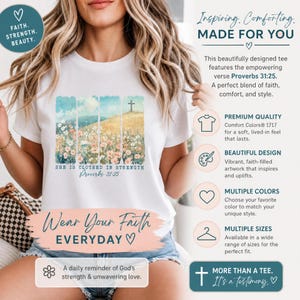 Puede incluir: Una camiseta blanca de manga corta con un estampado gráfico de un campo de flores silvestres bajo un cielo azul con una cruz en una colina. El texto "SHE IS CLOTHED IN STRENGTH" y "Proverbs 31:25" aparece en letras azules bajo la ilustración.