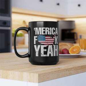 May include: A black ceramic mug featuring the white distressed text 'MERICA F*CK YEAH' with a graphic of the United States map filled with the American flag pattern. The design is centered on the front of the mug, which has a curved handle.