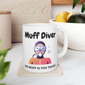 May include: White ceramic mug with the text "Muff Diver" and an illustration of a scuba diver. The diver graphic is wearing goggles and a breathing apparatus. The bottom of the mug reads "NO MUFF IS TOO TOUGH".