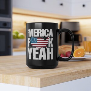 May include: A black ceramic mug featuring the white text 'MERICA F K YEAH' with a graphic of the United States map filled with the American flag pattern. The design is centered on the front of the mug, which has a curved handle on the right side.