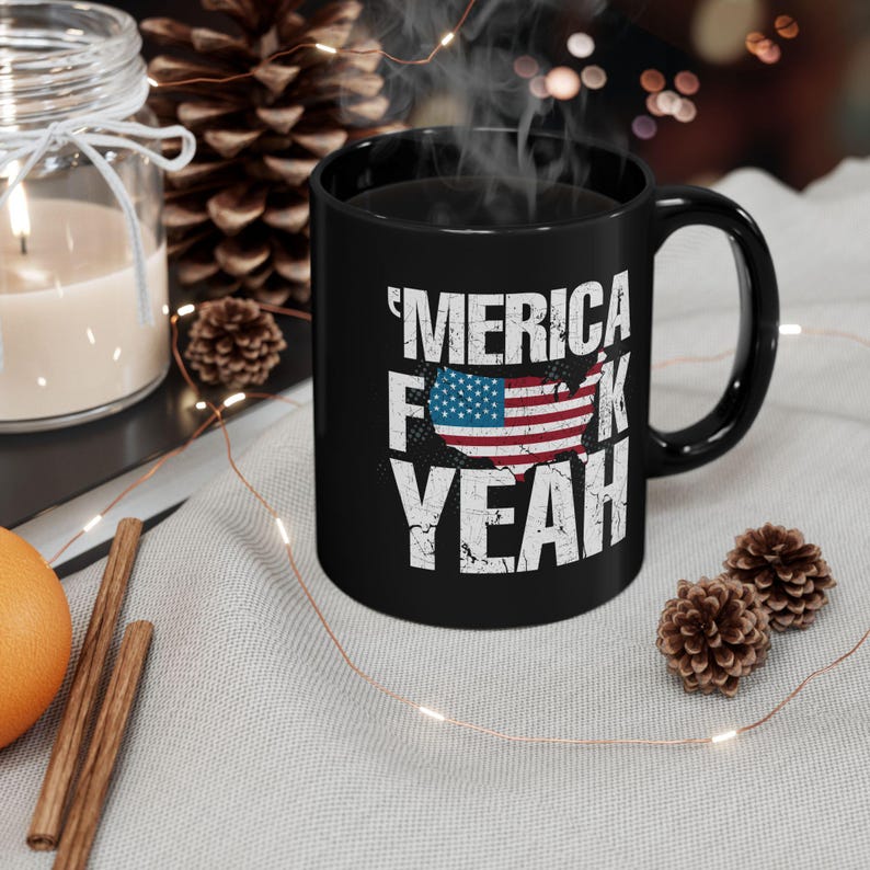 May include: A black ceramic mug featuring the white distressed text 'MERICA F K YEAH with a graphic of the United States map filled with the American flag pattern. The mug has a standard handle and is filled with a hot beverage emitting visible steam.