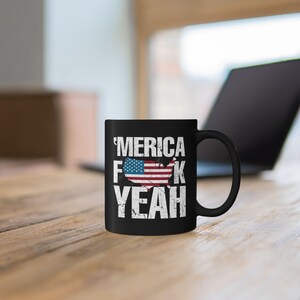 May include: A black ceramic mug featuring the white distressed text 'MERICA F*CK YEAH with a graphic of the United States map filled with the American flag pattern. The design is centered on the front of the mug, which has a standard handle.