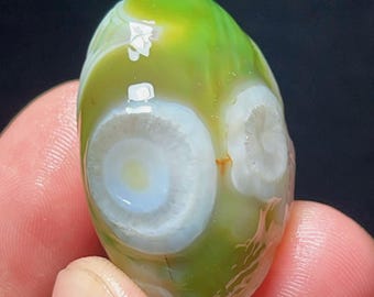 May include: A polished, oval-shaped green agate gemstone featuring two distinct white, circular eye-like patterns. The stone displays translucent lime and olive green hues with a smooth, glossy surface that reflects light, highlighting the natural banding.