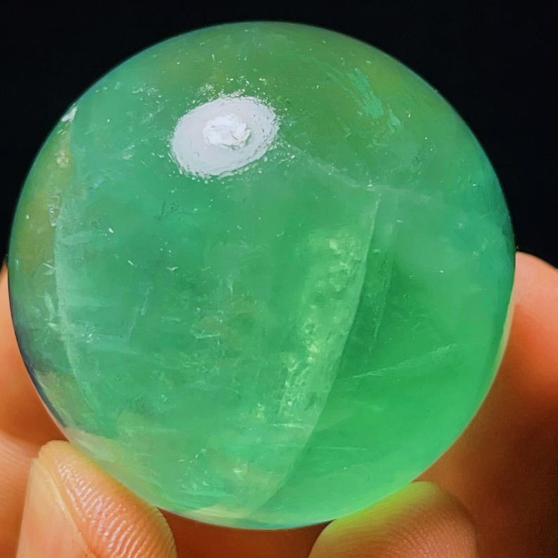 May include: A smooth, green fluorite sphere with a white, cloudy inclusion. The sphere is polished and has a translucent appearance.