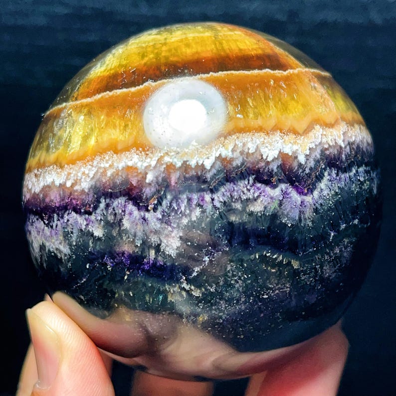 May include: A spherical gemstone with layers of yellow, orange, white, purple, and dark blue. The sphere features a bright, white, circular inclusion. The gemstone is held in a hand.
