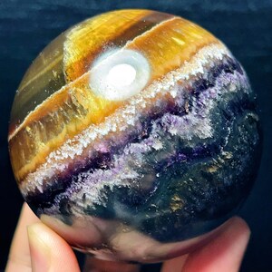 May include: A spherical gemstone, polished to a smooth finish, displaying bands of yellow, brown, purple, and white. The crystal ball has a bright, white, circular inclusion. The gemstone is held in a hand.