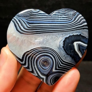 May include: Heart-shaped stone with a polished surface. The stone features a pattern of concentric circles and bands in shades of black, white, and blue. The heart is held in a hand, showcasing the natural stone's unique design.