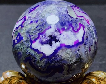 May include: A spherical gemstone, showcasing a vibrant mix of colours. The orb presents deep purple, white, and green hues, with complex patterns and a geode-like cavity. It is displayed on a decorative gold-coloured stand.