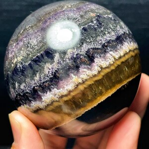 May include: A spherical crystal, displaying layers of purple, gold, and white. The polished surface reflects light, highlighting the crystal's internal patterns. The sphere is held in a hand, emphasizing its size and form.