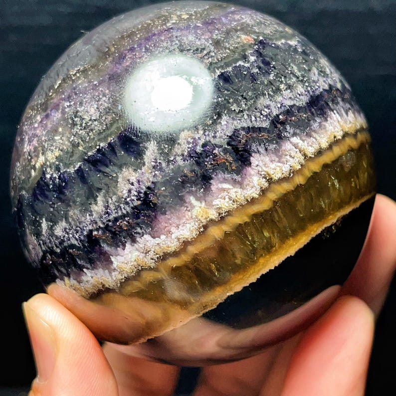 May include: A spherical crystal, displaying layers of purple, gold, and white. The polished surface reflects light, highlighting the crystal's internal patterns. The sphere is held in a hand, emphasizing its size and form.