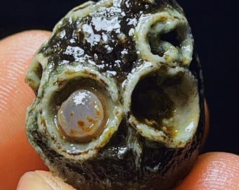 May include: A unique, naturally formed rock with a skull-like appearance. The rock features three circular openings, resembling eye sockets and a nose, with a central, light-colored stone in one opening. The rock's surface is a mix of dark brown, green, and white.