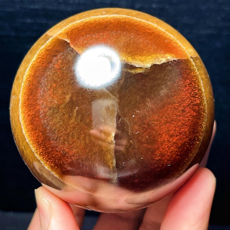 May include: A spherical gemstone with a polished surface, displaying a warm colour palette. The orb features amber, brown, and reddish-orange hues, with a bright, reflective spot. The stone is held in a hand, showcasing its size and texture.