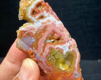 May include: A polished agate specimen featuring intricate banding in shades of orange, red, and white. The surface displays translucent botryoidal formations and circular patterns throughout the stone, showcasing a natural layered structure.