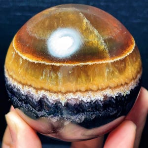 May include: A spherical gemstone with bands of colour. The orb features layers of brown, yellow, and black, with a white, eye-like formation at the top. The stone is held in a hand, showcasing its size and detail.