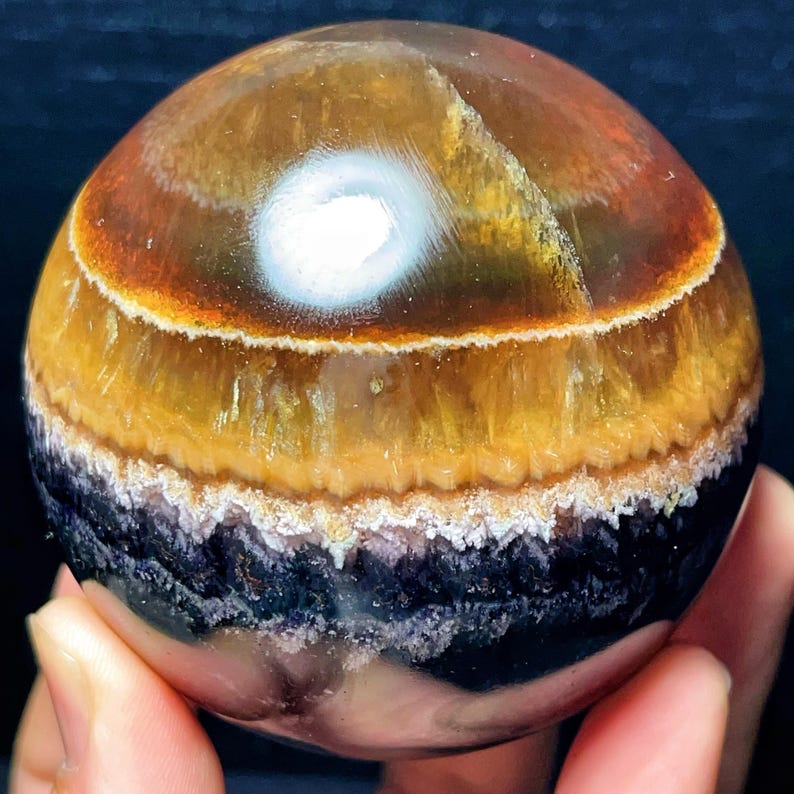 May include: A spherical gemstone with bands of colour. The orb features layers of brown, yellow, and black, with a white, eye-like formation at the top. The stone is held in a hand, showcasing its size and detail.