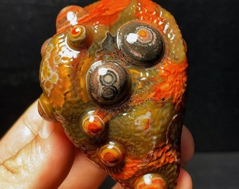 May include: A polished, multi-colored agate stone with a unique, eye-like pattern. The stone is a vibrant mix of orange, red, brown, and yellow.