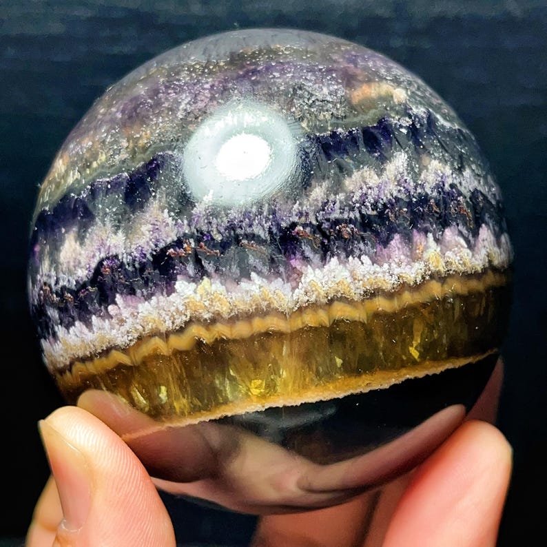 May include: A crystal sphere with layers of purple, white, and gold. The sphere is held in a hand, highlighting its polished surface. The crystal's bands create a unique pattern. The sphere is a decorative item, with a diameter of approximately 7 cm.